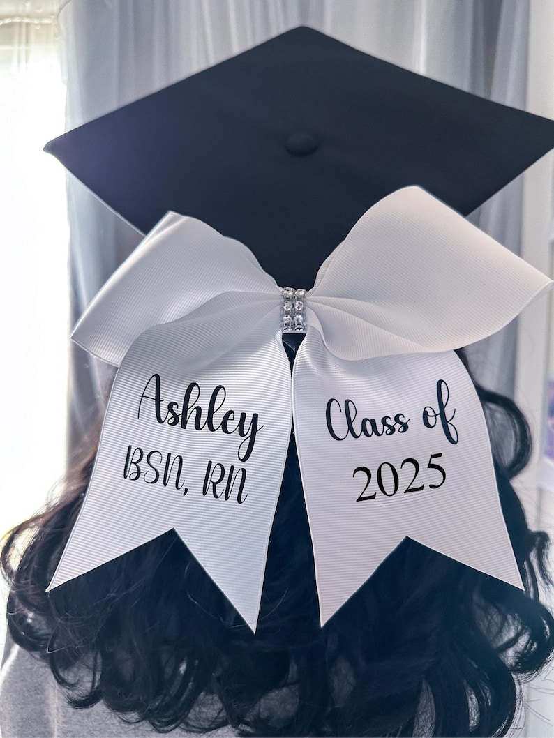Peut inclure: Noeud de graduation blanc avec du texte noir qui dit "Ashley BSN, RN" d'un c&ocirc;t&eacute; et "Class of 2025" de l'autre. Le n&oelig;ud est d&eacute;cor&eacute; d'un embellissement en strass.