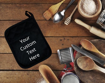 May include: Black oven mitt with white text that reads "Your Custom Text Here" on a wooden table with various kitchen utensils, including spoons, a rolling pin, and a grater.