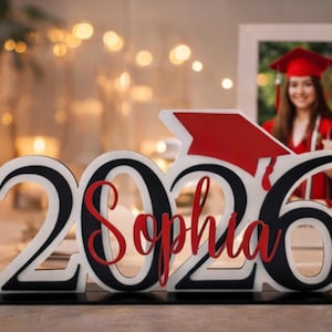 May include: A graduation centerpiece with the year "2026" and the name "Sophia" in red script. The numbers are white with black outlines, and a red graduation cap is attached. A framed photo of a graduate in a red gown is in the background.