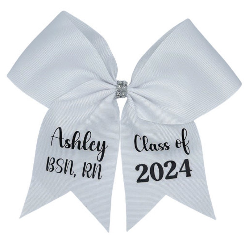 Peut inclure: Un n&oelig;ud de cheerleading blanc avec un strass argent&eacute; au centre. Le c&ocirc;t&eacute; gauche du n&oelig;ud porte le texte "Ashley BSN, RN" et le c&ocirc;t&eacute; droit porte le texte "Class of 2024".