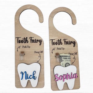 May include: Two wooden tooth fairy door hangers with a tooth cutout, one with a blue name "Nick" and the other with a pink name "Sophia". Each hanger has a small jar for a tooth and a space for money. The text "Tooth Fairy" and "Pick Up" and "Drop Off" are written on each hanger.