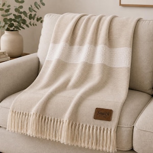 Puede incluir: Una manta de punto en color beige y crema con rayas horizontales acanaladas y una banda ancha texturizada. La manta tiene flecos largos en el borde inferior y un parche rectangular de cuero marrón con el texto Smith EST 2023 cosido.
