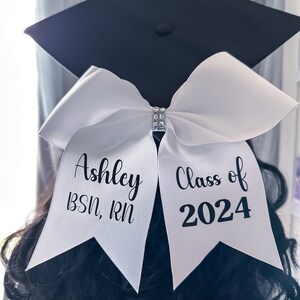 Peut inclure: Chapeau et robe de graduation blancs avec un n&oelig;ud blanc et le texte "Ashley BSN, RN Classe de 2024" dessus.