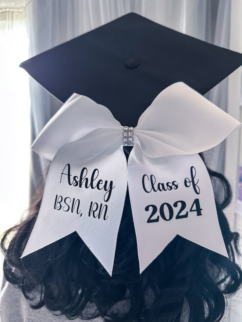Peut inclure: Chapeau et robe de graduation blancs avec un n&oelig;ud blanc et le texte "Ashley BSN, RN Classe de 2024" dessus.