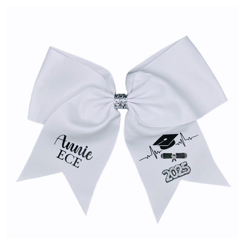 Peut inclure: Noeud de graduation blanc avec un bonnet de graduation noir, un dipl&ocirc;me et un graphique de battement de c&oelig;ur. Le n&oelig;ud porte &eacute;galement le texte "Annie ECE 2025" imprim&eacute; dessus.