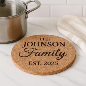May include: A round cork trivet with the text "THE JOHNSON Family EST. 2025" in a dark brown color. A stainless steel pot and a striped kitchen towel are in the background. The trivet is designed to protect surfaces from heat.