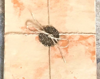 Op de afbeelding: Een stapel briefpapier met een aquarel-achtig ontwerp in perzik- en oranjetinten. Het papier is vastgebonden met touw en verzegeld met een decoratief, donkergekleurd versiering. De algehele esthetiek is rustiek en artistiek.