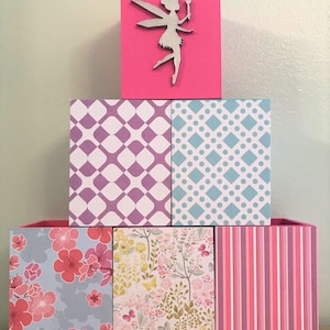 Puede incluir: Una pila de cajas de almacenamiento decorativas en varios patrones y colores. La caja superior es rosa intenso con una silueta de hada plateada. Las otras cajas presentan diseños florales, geométricos y a rayas en tonos de rosa, morado y azul.