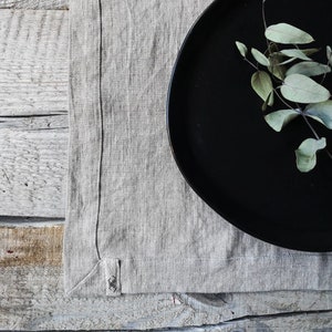 Puede incluir: Un plato negro con una rama de hojas verdes se encuentra sobre un mantel individual de lino beige. El mantel individual tiene un borde cosido y un pequeño diseño bordado en la esquina.