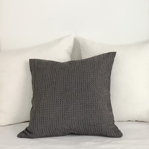 Può includere: Un letto con biancheria da letto in lino bianco e un cuscino di lancio in tessuto grigio. La testiera è realizzata con travi in legno a vista. Un arazzo da parete intrecciato è sopra il letto.