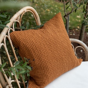 Könnte beinhalten: Ein braunes, gewaffelt strukturiertes Kissen auf einem weißen Korbstuhl. Der Stuhl steht im Freien in einem Garten.