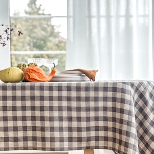 Puede incluir: Un mantel a cuadros grises y beige cubre una mesa de madera, decorada con calabazas decorativas y platos. La mesa está situada cerca de una ventana con vistas a árboles y cortinas blancas. El mantel tiene un patrón de cuadros clásico.