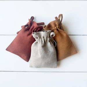 Puede incluir: Tres pequeñas bolsas con cordón en tonos óxido, beige y gris. Las bolsas están hechas de una tela texturizada y tienen una parte superior fruncida con un lazo para cerrar. El fondo es una superficie de madera blanca.