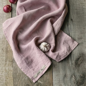 Op de afbeelding: Een roze linnen keukendoek met een wit label met de tekst "JAUJA" op een houten oppervlak. Er liggen drie rode uien en een teentje knoflook op de doek.
