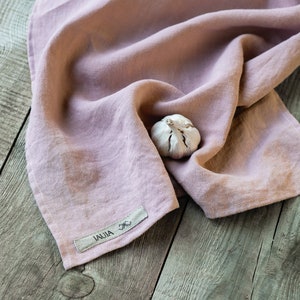 May include: A dusty rose linen kitchen towel with a small white tag that says "IAUJA" on a wooden surface. A single garlic clove is resting on the towel.