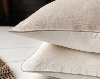 May include: Two linen pillow covers, one beige and one white, with a subtle brown trim around the edges. The pillows are stacked on top of each other on a white wooden surface.