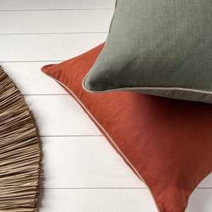 May include: Two decorative throw pillows with a white trim. The top pillow is a muted green color and the bottom pillow is a burnt orange color.