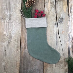 Peut inclure: Une chaussette de Noël en lin vert avec un revers beige, remplie de baies rouges et d'une pomme de pin.
