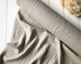 May include: A roll of beige fabric with a textured, crinkled appearance. The fabric is draped over a white surface, with a portion rolled up. Dried flowers are visible in the background.