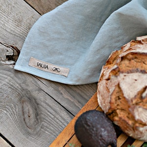 Op de afbeelding: Een lichtblauwe linnen keukendoek met een bruin label waarop "JAUJA" en een klein ontwerp staan. De handdoek is gevouwen en ligt op een houten oppervlak met een brood en een houten snijplank.