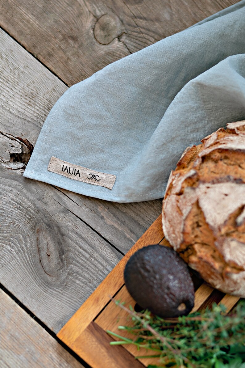 Op de afbeelding: Een lichtblauwe linnen keukendoek met een bruin label waarop "JAUJA" en een klein ontwerp staan. De handdoek is gevouwen en ligt op een houten oppervlak met een brood en een houten snijplank.