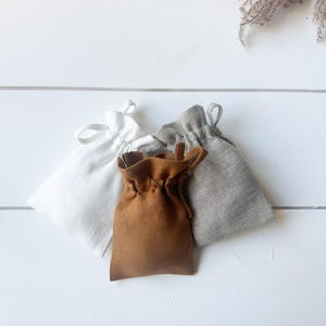 Puede incluir: Tres peque&ntilde;as bolsas con cord&oacute;n en blanco, marr&oacute;n y gris, dispuestas sobre una superficie de madera blanca. Las bolsas son de tela y tienen un dise&ntilde;o sencillo y minimalista. Una ramita de flores secas est&aacute; en la esquina superior derecha.