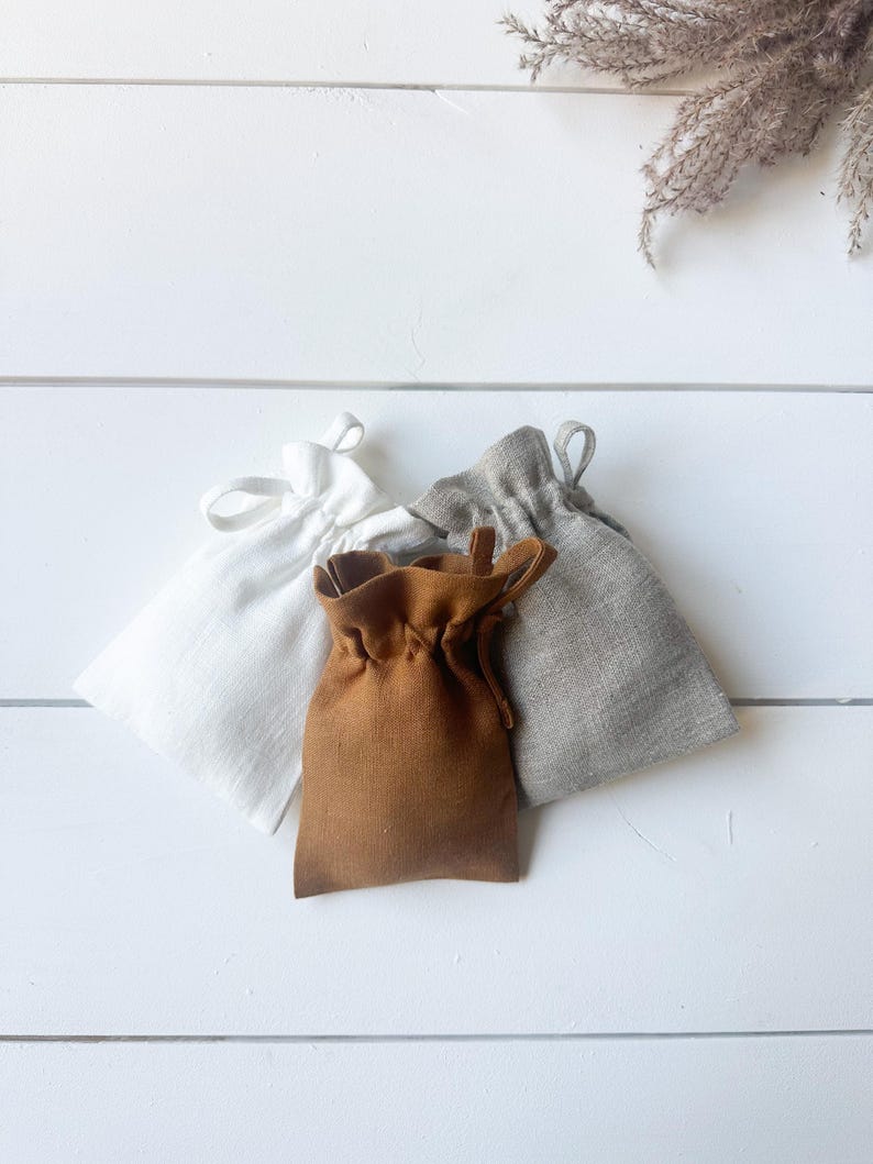 Puede incluir: Tres peque&ntilde;as bolsas con cord&oacute;n en blanco, marr&oacute;n y gris, dispuestas sobre una superficie de madera blanca. Las bolsas son de tela y tienen un dise&ntilde;o sencillo y minimalista. Una ramita de flores secas est&aacute; en la esquina superior derecha.