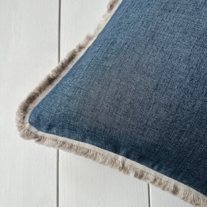 May include: Close-up of a square denim blue pillow with a fringed edge. The pillow has a light beige fringe and a light beige border. The pillow is on a white surface.