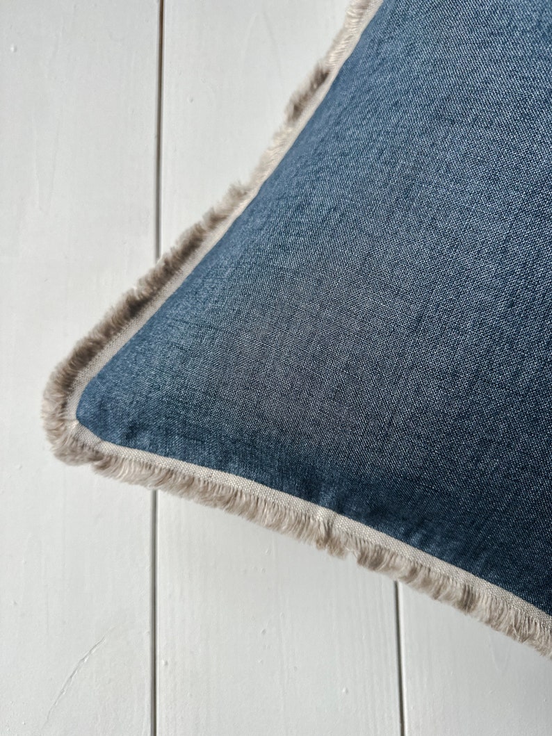May include: Close-up of a square denim blue pillow with a fringed edge. The pillow has a light beige fringe and a light beige border. The pillow is on a white surface.