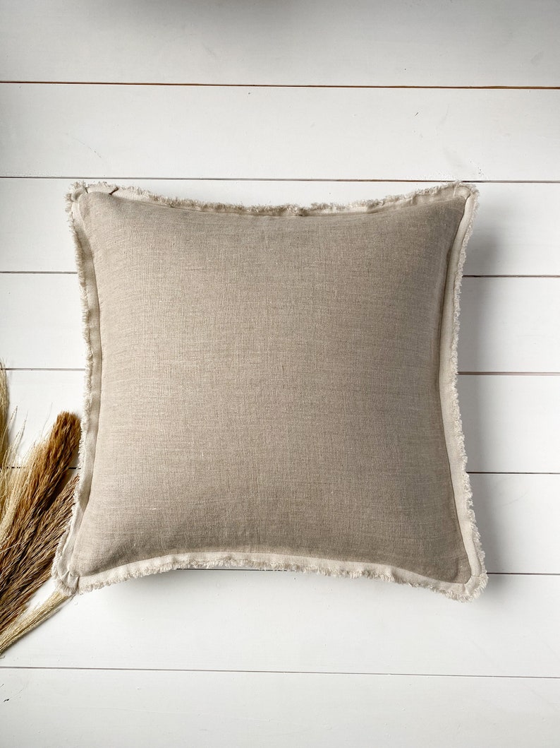 May include: A square beige linen pillow with a fringed edge. The pillow is centered on a white wooden surface, with dried ornamental grass in the lower left corner. The pillow's neutral color and texture suggest a home decor item.