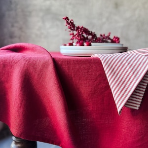 Puede incluir: Un mantel de lino rojo con una servilleta a rayas blancas y rojas encima. Un plato blanco con bayas rojas está sobre la mesa.