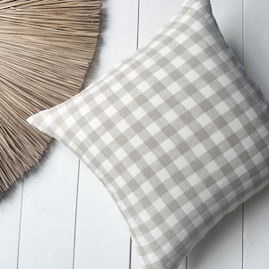 Puede incluir: Una almohada cuadrada con un patrón de cuadros beige y blanco. La almohada está hecha de lino y tiene una textura suave.