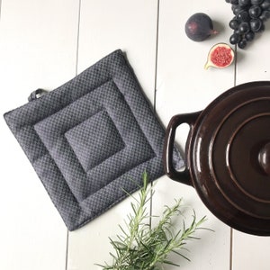 Peut inclure: Un dessous de plat matelassé gris de forme carrée. Le dessous de plat est posé sur une surface en bois blanc avec une casserole brune, des brins de romarin et des figues.