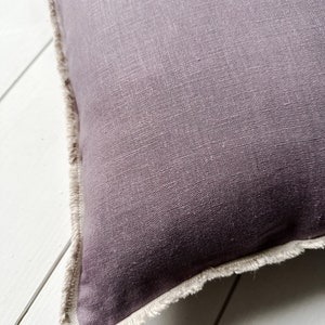 May include: Close-up of a square, plum-colored pillow with a fringed edge. The pillow has a textured linen appearance. The fringed edge is a light beige color, creating a contrast with the darker pillow.