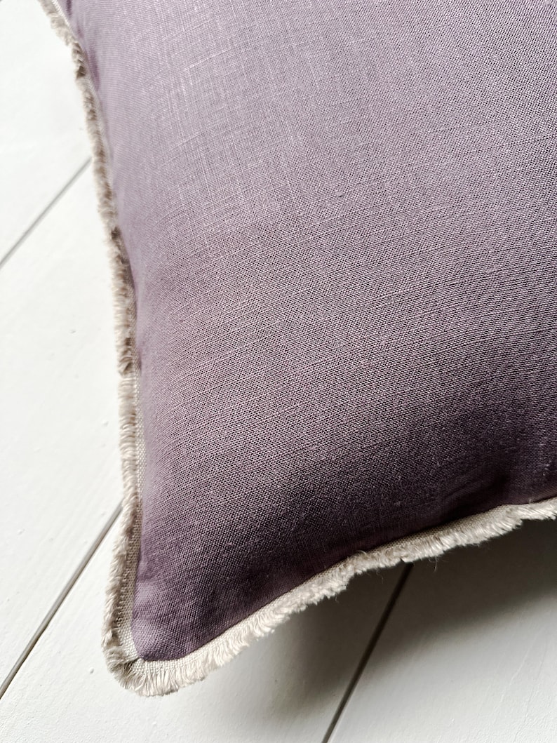 May include: Close-up of a square, plum-colored pillow with a fringed edge. The pillow has a textured linen appearance. The fringed edge is a light beige color, creating a contrast with the darker pillow.