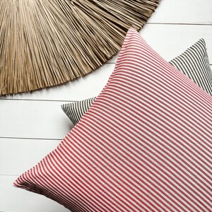 May include: Two striped cushion covers, one red and white, and one black and white, on a white wood background. A large, round, woven straw wall hanging is above the cushions.