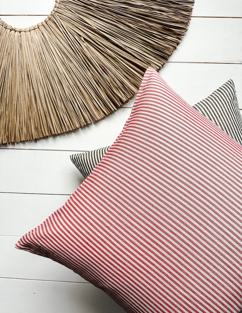 May include: Two striped cushion covers, one red and white, and one black and white, on a white wood background. A large, round, woven straw wall hanging is above the cushions.