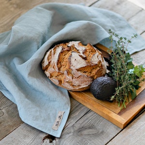 Op de afbeelding: Een rustiek houten snijplank met een zuurdesembrood, een avocado en takjes verse kruiden. De plank is bedekt met een lichtblauwe linnen theedoek met de tekst "HUA JC" erop gedrukt.