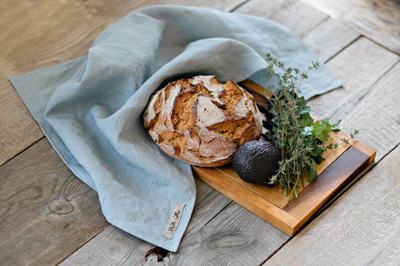 Op de afbeelding: Een rustiek houten snijplank met een zuurdesembrood, een avocado en takjes verse kruiden. De plank is bedekt met een lichtblauwe linnen theedoek met de tekst "HUA JC" erop gedrukt.