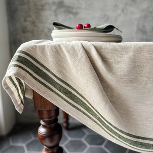 K&ouml;nnte beinhalten: Eine beige Tischdecke aus Leinen mit drei gr&uuml;nen Streifen am Rand. Die Tischdecke ist &uuml;ber einen Holztisch mit einem gedrehten Bein gelegt. Der Tisch steht auf einem grauen sechseckigen Fliesenboden.