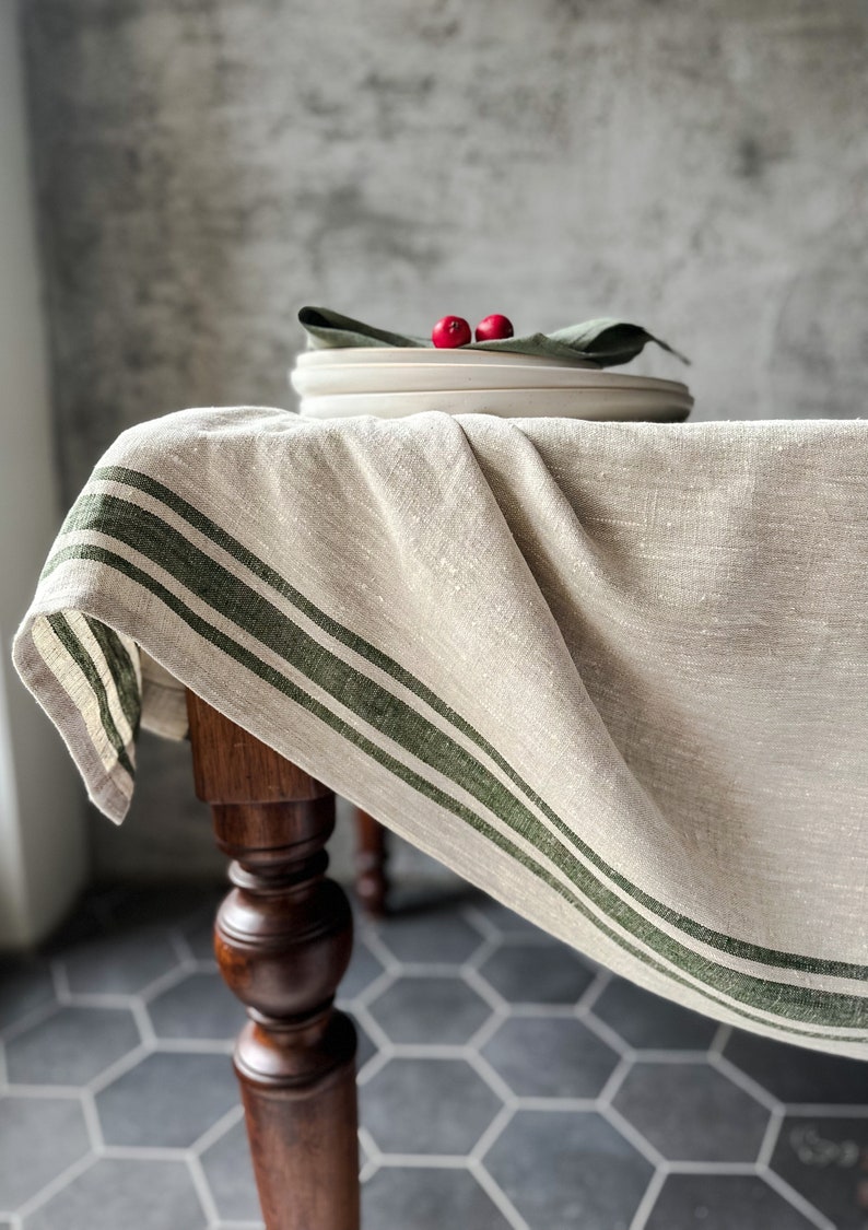 K&ouml;nnte beinhalten: Eine beige Tischdecke aus Leinen mit drei gr&uuml;nen Streifen am Rand. Die Tischdecke ist &uuml;ber einen Holztisch mit einem gedrehten Bein gelegt. Der Tisch steht auf einem grauen sechseckigen Fliesenboden.