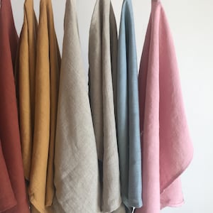 May include: Seven linen tea towels hanging on a white rod. The towels are in various colours, including rust, mustard yellow, natural linen, light blue, and pink.