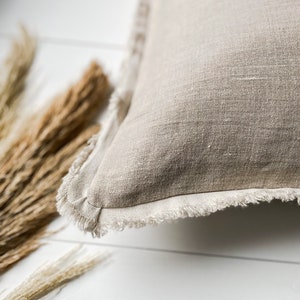 May include: Close-up of a beige linen pillow with a fringed edge. The pillow is square-shaped and has a textured surface. Dried wheat stalks are in the background, resting on a white surface.