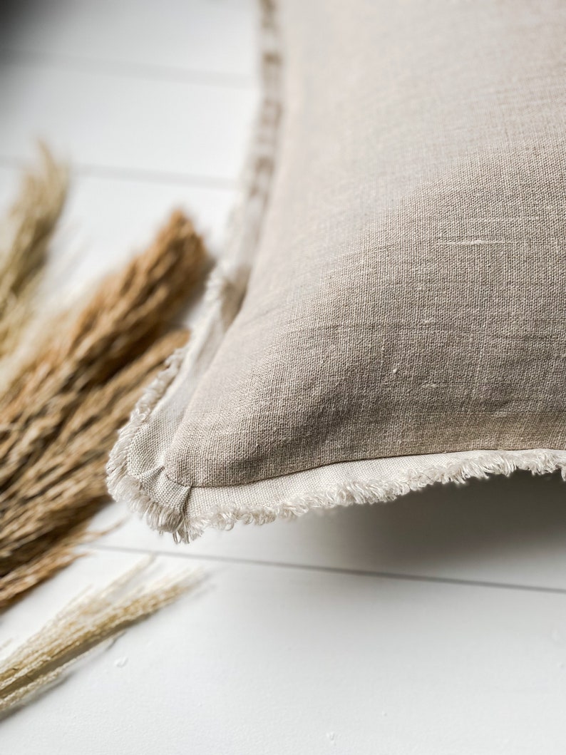 May include: Close-up of a beige linen pillow with a fringed edge. The pillow is square-shaped and has a textured surface. Dried wheat stalks are in the background, resting on a white surface.