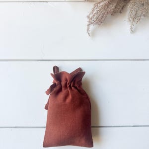 Puede incluir: Una peque&ntilde;a bolsa con cord&oacute;n de color &oacute;xido hecha de tela texturizada. La bolsa es rectangular con una parte superior fruncida y un peque&ntilde;o lazo para cerrar. El fondo es una superficie de madera blanca con hierba ornamental seca.