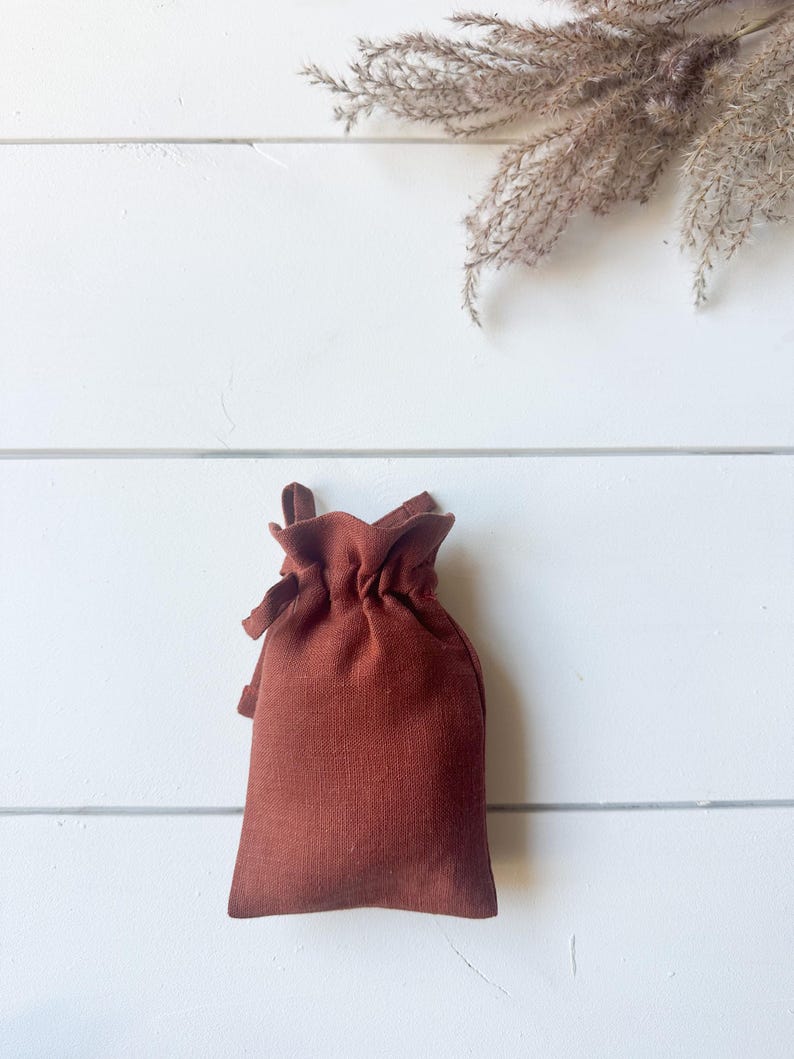 Puede incluir: Una peque&ntilde;a bolsa con cord&oacute;n de color &oacute;xido hecha de tela texturizada. La bolsa es rectangular con una parte superior fruncida y un peque&ntilde;o lazo para cerrar. El fondo es una superficie de madera blanca con hierba ornamental seca.
