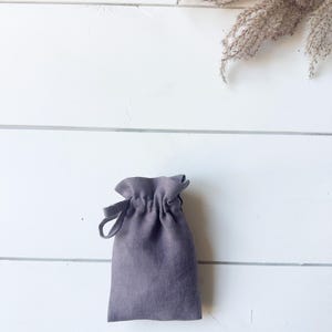 Puede incluir: Una peque&ntilde;a bolsa gris con cord&oacute;n hecha de tela texturizada. La bolsa est&aacute; fruncida en la parte superior con un cierre de cord&oacute;n y tiene un lazo en el lateral. El fondo es una superficie de madera blanca con flores secas.