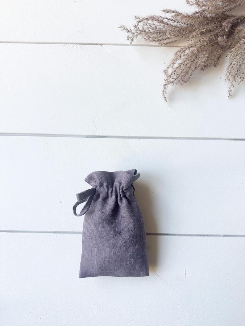 Puede incluir: Una peque&ntilde;a bolsa gris con cord&oacute;n hecha de tela texturizada. La bolsa est&aacute; fruncida en la parte superior con un cierre de cord&oacute;n y tiene un lazo en el lateral. El fondo es una superficie de madera blanca con flores secas.