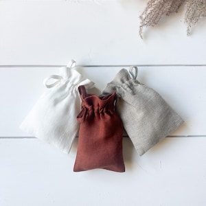 Puede incluir: Tres peque&ntilde;as bolsas con cord&oacute;n en blanco, &oacute;xido y gris, dispuestas sobre una superficie de madera blanca. Las bolsas est&aacute;n hechas de una tela texturizada, con una parte superior fruncida y un cierre de cord&oacute;n a juego. Hierba ornamental seca en el fondo.