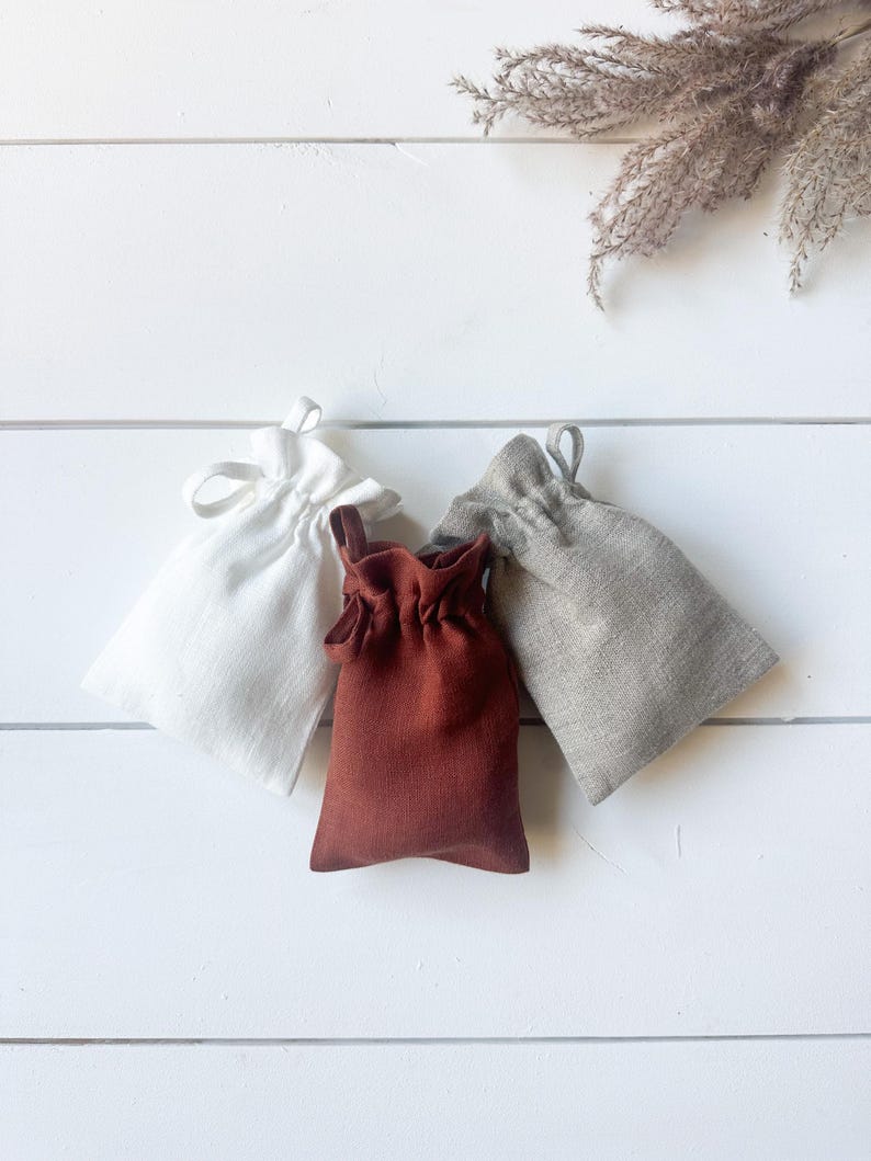 Puede incluir: Tres peque&ntilde;as bolsas con cord&oacute;n en blanco, &oacute;xido y gris, dispuestas sobre una superficie de madera blanca. Las bolsas est&aacute;n hechas de una tela texturizada, con una parte superior fruncida y un cierre de cord&oacute;n a juego. Hierba ornamental seca en el fondo.